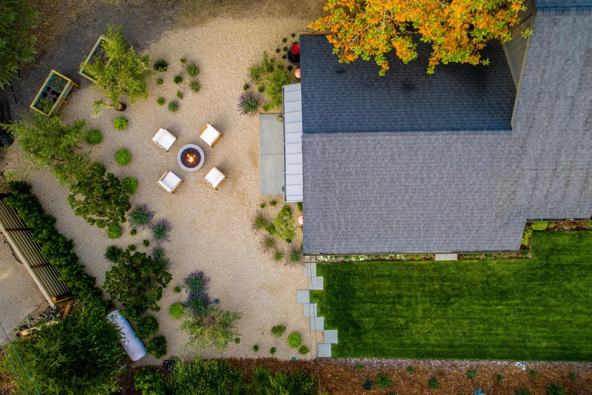 Aerial view of a beautifully landscaped backyard featuring a fire pit seating area, gravel pathways, lush greenery, and a well-maintained lawn.